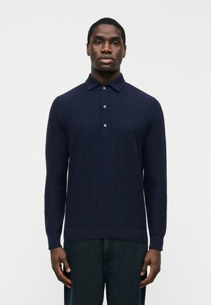 Hombre con polo azul marino ajustado de manga larga acanalada con tres botones, de pie derecho contra un fondo liso y claro.