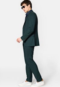 Ciemnozielony garnitur z gładkiego materiału, połączony z granatową koszulą w kropki i białymi trampkami. Noszone okulary przeciwsłoneczne; schludny, dopasowany krój.