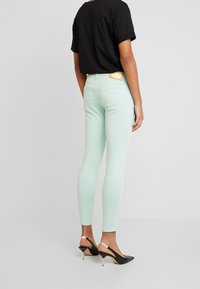 Skinny Jeans in Mintgrün mit glatter Textur, ausgestattet mit Gesäßtaschen und einem goldenem Logo-Patch. Kombiniert mit einem schwarzen T-Shirt und schwarzen High Heels.
