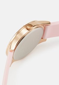 Rose gold metallisk klokke med rund kasse, rosa silikonrem og teksturert krone. Glatt solid bakside uten synlige merker.