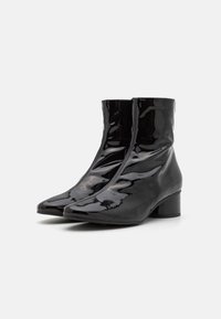 Botines de charol negros con punta afilada, cremallera lateral y tacón grueso. La textura suave y el diseño elegante crean un aspecto moderno.