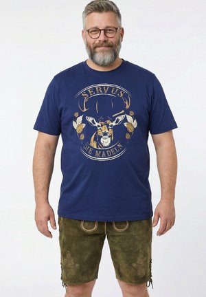 Mann mit grauem Bart und Brille, der ein marineblaues T-Shirt mit einem Hirschmotiv und den Worten „Servus Die Madeln“ sowie grüne bestickte Shorts trägt.