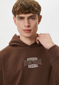 Brun hættetrøje med en kængurulomme, der har hvid broderet tekst "SUPERIOR NEW YORK ORIGINAL". Blødt stof med struktureret finish.