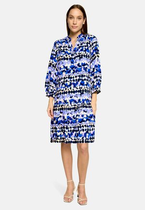 Vrouw draagt een knielange, loszittende jurk met blauwe, zwarte en witte cirkelpatronen en driekwartmouwen, staand in neutrale schoenen.