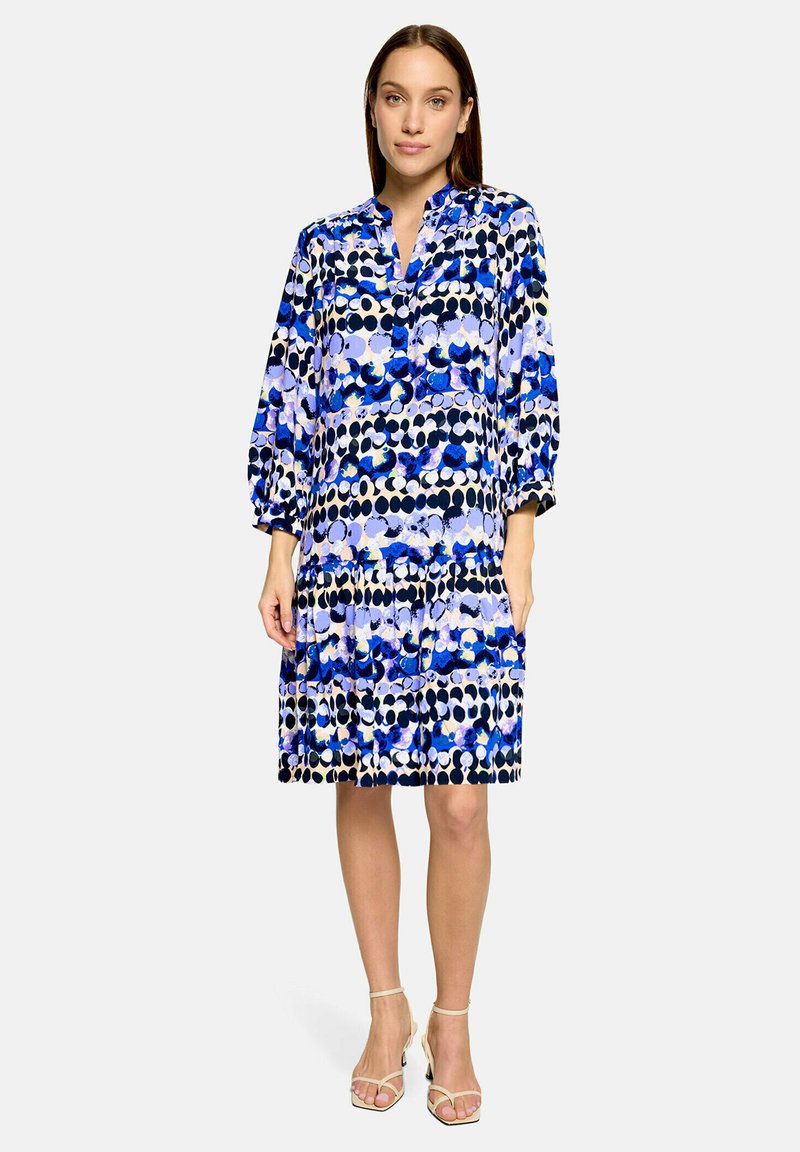 Femme portant une robe ample mi-longue avec des motifs circulaires bleus, noirs et blancs et des manches trois-quarts, debout avec des chaussures neutres.