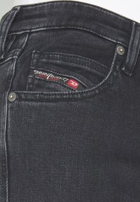 Svarta denimjeans med design på bakfickorna, synlig sömnad och en röd och vit kärnmärke. Metallknappstängning är närvarande.
