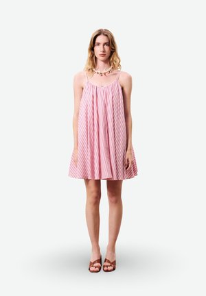 Jeune femme debout portant une robe rose à rayures sans manches, des talons ouverts marron et un collier épais en perles, face à l'avant sur un fond blanc.