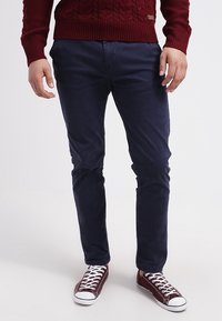 Mann trägt dunkelblaue, schmal geschnittene Hose, bordeauxfarbenen Strickpullover und passende bordeauxfarbene Leinenschuhe mit weißen Schnürsenkeln und Sohlen, steht auf weißem Boden.