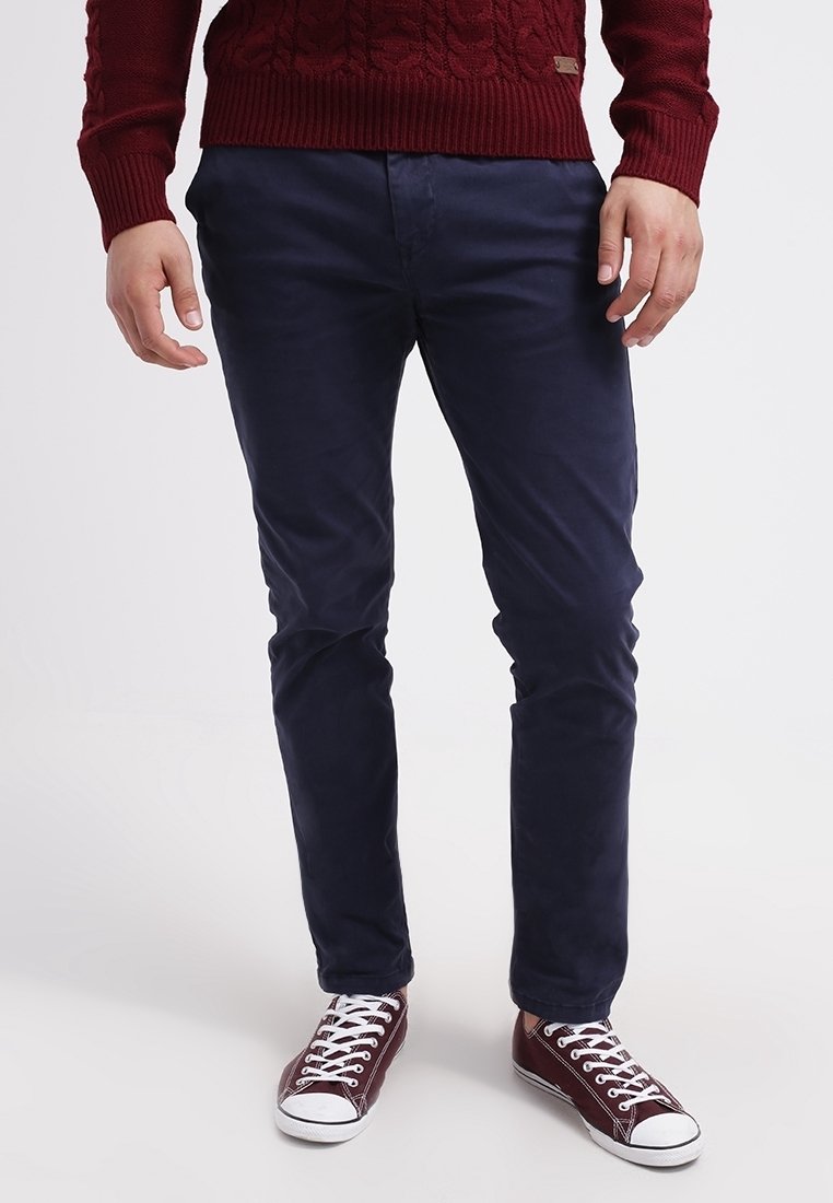 Mann trägt dunkelblaue, schmal geschnittene Hose, bordeauxfarbenen Strickpullover und passende bordeauxfarbene Leinenschuhe mit weißen Schnürsenkeln und Sohlen, steht auf weißem Boden.