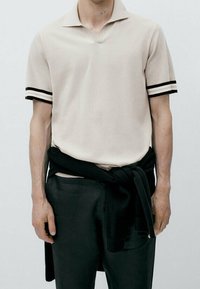 Polo-shirt beige à manches courtes avec des accents de rayures noires, assorti à un pantalon noir et un pull noir noué autour de la taille.
