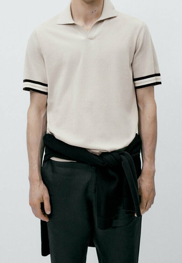 Polo-shirt beige à manches courtes avec des accents de rayures noires, assorti à un pantalon noir et un pull noir noué autour de la taille.