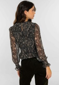 Top en negro y beige con estampado, mangas con volantes y detalle de abertura en la espalda. Hecho de una tela ligera y transparente. Combinado con pantalones verde oscuro.