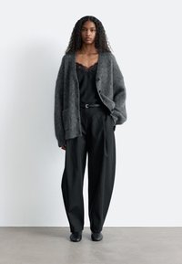 Graue, flauschige Strickjacke mit Knöpfen vorne und Seitentaschen, darüber ein schwarzes Spitzenoberteil und weit geschnittene schwarze Hosen. Schwarze Schuhe.