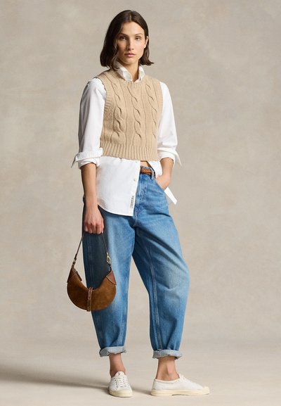 Žena má na sebe béžovú kabelkovanú vestu cez bielu košeľu, modré vyhrnuté rifle, biele tenisky a drží malú hnedú kabelku.