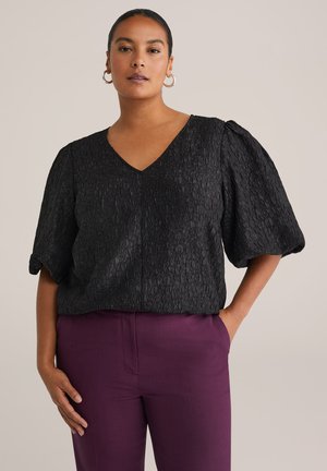 Blusa nera con texture, scollatura a V e maniche a sbuffo, abbinata a pantaloni viola, che mostrano un materiale liscio e una vestibilità comoda.