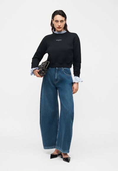 Femme portant un sweat-shirt noir, un chemisier bleu clair avec des poignets et un col à volants, un jean bleu à jambes larges, des chaussures noires à bouts pointus, et tenant une pochette cloutée.