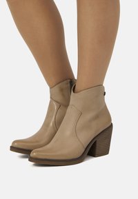 Botines de cuero beige hasta el tobillo con puntera puntiaguda, cremallera lateral y un tacón de madera apilada. Costuras decorativas a lo largo de las costuras.