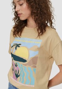 Beigefarbenes Baumwoll-T-Shirt mit einem bunten Grafikdesign eines Sonnen, einer Palme und Händen über abstrakten Wellen, mit dem Text "Grüße von der Sonnenseite."