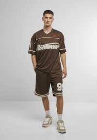 Tenue de sport marron comprenant un maillot à manches courtes avec des accents blancs et du texte, associé à un short assorti et des baskets légères.