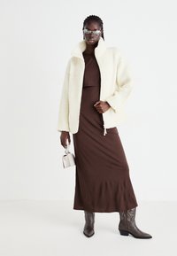 Giacca in shearling crema con colletto alto, vestito aderente marrone, stivali da cowboy grigi e una piccola borsa off-white. Design e textures minimalisti.