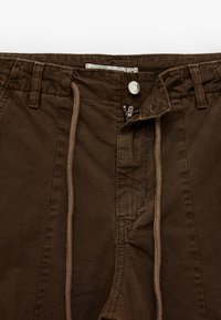 Brązowe bawełniane szorty z zapięciem na zamek i guzik, sznurkiem w pasie oraz kieszeniami po bokach. Gładka tekstura z przeszyciami.