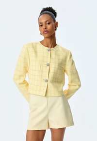 Giacca gialla in tweed con motivo testurizzato, caratterizzata da un colletto rotondo, maniche lunghe e bottoni decorativi argentati. Abbinata a pantaloni corti gialli.