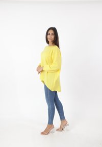 Gelber, übergroßer Strickpullover mit abgerundetem Saum, getragen über eng anliegenden blauen Jeans, kombiniert mit transparenten Absatzsandalen. Glatte Textur.