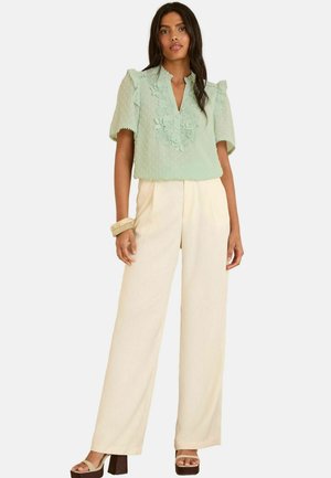 Blusa verde menta con ricami floreali e maniche a volant, abbinata a pantaloni a gamba larga color crema e tacchi chunky. Tessuto dalla consistenza liscia.