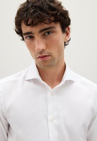 Giovane uomo con capelli ricci e scuri che indossa una camicia bianca con bottoni, posato davanti a uno sfondo chiaro, mentre guarda direttamente nella fotocamera.