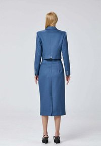 Ensemble blazer ajusté bleu et jupe crayon, fabriqué dans un tissu lisse, présentant un design cropped, des manches longues et une fente au dos de la jupe.