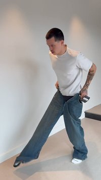 Man with tattoos wearing oversized jeans and white t-shirt adjusts pants while standing in a minimal indoor space with light walls.