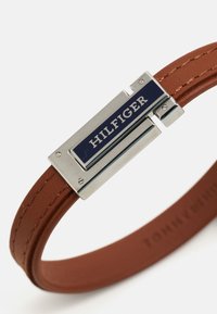 Pruun nahkavööstik, millel on hõbedane metalllukk, millel on sinine ja valge "HILFIGER" logo plaat ning õmblusdetailid rihmal.