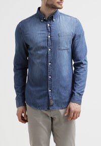 Camisa de mezclilla, azul medio, de mangas largas, con cuello de botón, un bolsillo en el pecho, botones blancos, dobladillo curvado, textura suave.