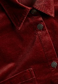 Lähellä punainen corduroy-paita, jossa on teksturoitu pinta, terävä kaulus, tummat metallinapit ja läpän tasku.