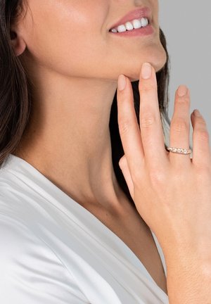 Gouden ring met drie heldere stenen, gezet op een slanke band. De hand van het model rust nabij de kin en toont de natuurlijke nagelkleur en de gladde huid.