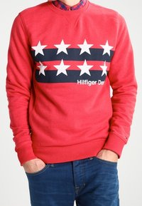 Sudadera roja con cuello redondo, mangas largas y un diseño frontal de estrellas alternas en negro y blanco, con el texto "Hilfiger Denim".