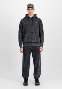Homme portant un sweat à capuche noir délavé avec un pantalon de jogging assorti, des baskets noires et une casquette de baseball grise, se tenant contre un fond neutre.