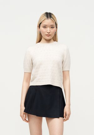 Jeune femme aux cheveux blonds raides porte un pull beige à manches courtes et une jupe noire plissée, debout devant un fond clair uni.