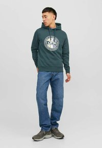Sudadera verde con un logo gráfico circular en el pecho, combinada con jeans azules y zapatillas grises. Ajuste simple y casual.
