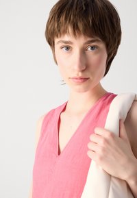 Roze halter haljina s dubokim V-izrezom, izrađena od teksturiranog materijala. Model drži krem boje odjevni predmet preko jednog ramena.