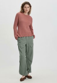 Rosenfarbener Rippstrickpullover kombiniert mit locker sitzenden olivgrünen Cargo-Hosen, die seitliche Taschen und eine entspannte Silhouette aufweisen. Braune Schuhe.