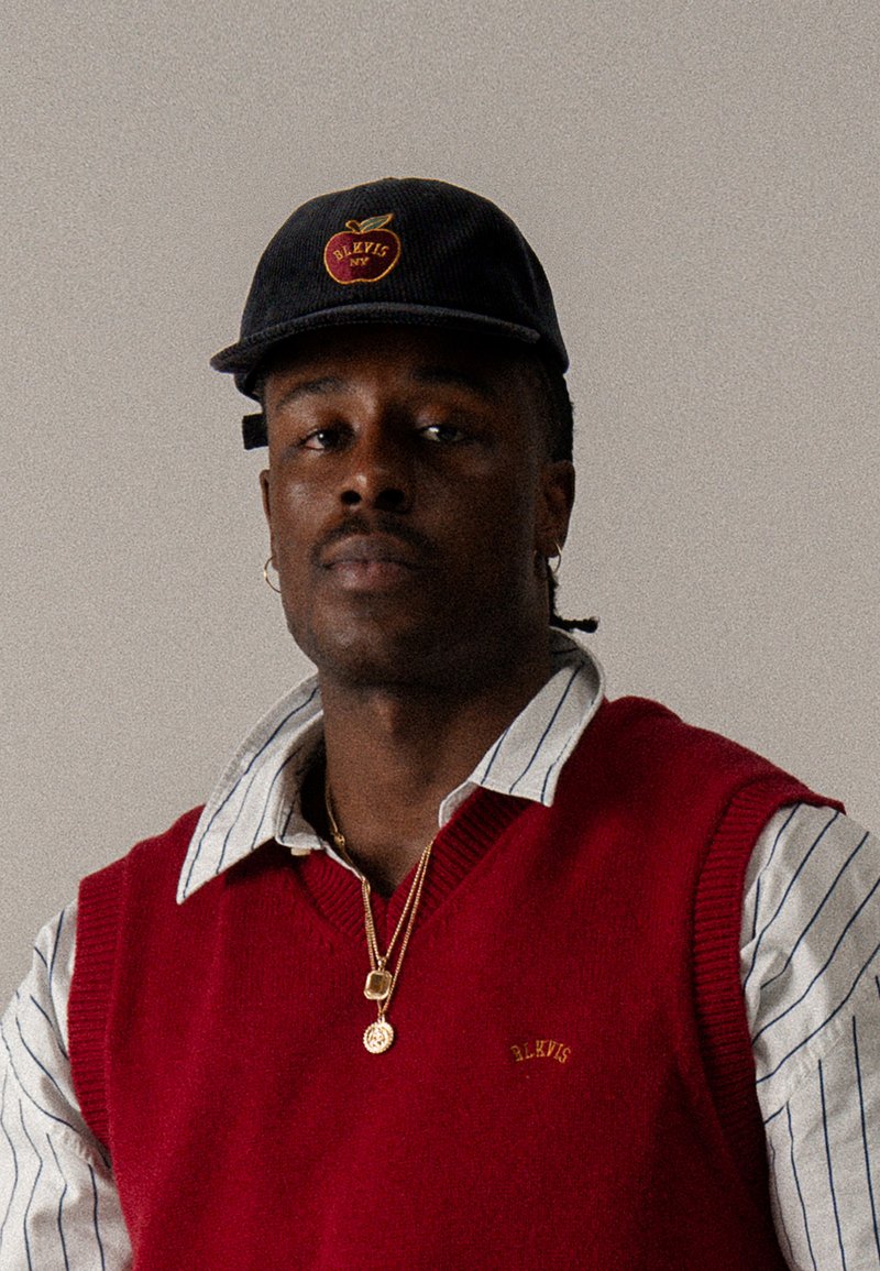 Chaleco de punto rojo sobre una camisa de cuello a rayas azul y blanca, combinado con una gorra oscura con el logo de una manzana y acentos dorados.