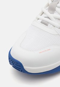 Fehér sportcipő texturált felsőrésszel és párnázott nyakkal. Kék gumi talpa van, amelyen piros színű "VÉDŐ ZÓNÁ" felirat található a lábujjak közelében.