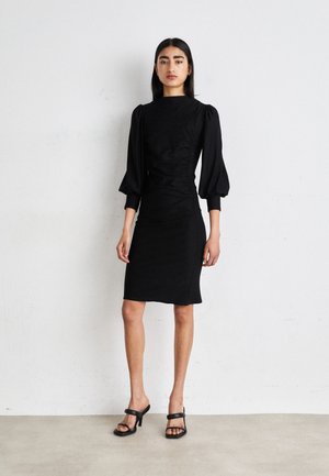 Black fitted dress with a high neck, puffed sleeves, and side ruching. Worn with black block-heeled sandals. Smooth fabric, sleek design.