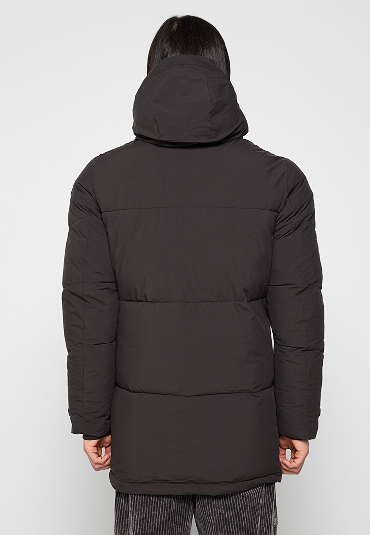 Personne portant un long manteau rembourré noir avec une capuche, vu de dos, se tenant contre un fond clair.