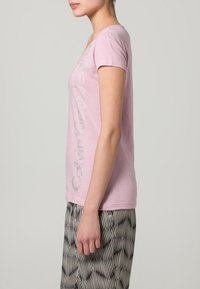 Camiseta de manga corta rosa con un diseño de texto brillante en el lado izquierdo; tejido suave con un corte relajado. Combinada con pantalones estampados.