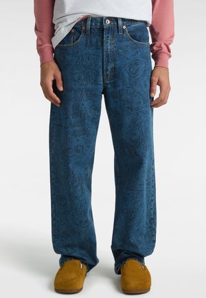 Widok z przodu osoby noszącej ciemnoniebieskie dżinsy z czarnymi nadrukami w stylu szkiców, musztardowożółte buty wsuwane, białą koszulkę i różową bluzę z długimi rękawami.