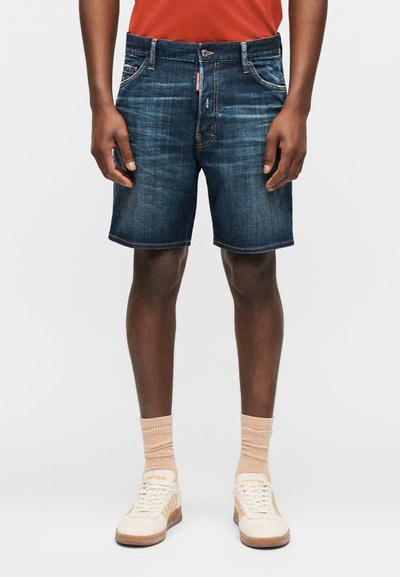 Άνδρας που φοράει σκούρα μπλε τζιν σορτς, μπεζ κάλτσες, λευκά αθλητικά παπούτσια και πορτοκαλί πουκάμισο, στέκεται με τα χέρια χαλαρά στα πλευρά.