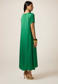 Maxi dress verde fluente con maniche corte arrotolate, texture liscia e vestibilità rilassata. Indossato con tacchi nudi e bracciali sovrapposti.