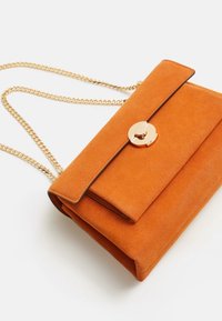 Sac à main en daim orange avec un fermoir circulaire doré et une bandoulière chaîne. Le sac possède un rabat de fermeture et une surface lisse et texturée.